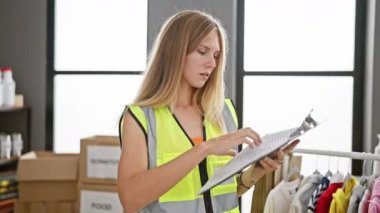 Yüksek görüş mesafesi olan sarışın bir kadın elbise askısı ve kutuları olan bir depoda envanter kontrolü yapıyor..