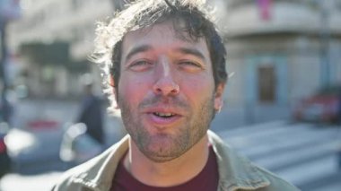 Güneşli şehir sokağında gülümseyen sakallı İspanyol adam bulanık şehir arka planıyla dostça ve rahat bir atmosfer taşıyor..