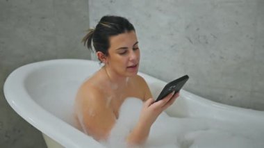 Rahat bir kadın, modern bir banyo ortamında köpük dolu bir küvette banyo yaparken akıllı telefon kullanır.