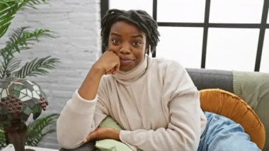 Ne mutlu bir başarı! Rasta saçlı genç siyahi bir kadın rahat bir koltukta oturuyor, baş parmak işareti yapıyor. Onaylamak ve evde mutlu görünmek.!