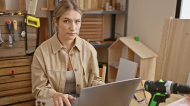 Marangozluk atölyesindeki genç, kendine güvenen sarışın kadın. Elinde bilgisayarla dur levhasını kaldırırken yüzünde ciddi bir ifade var.