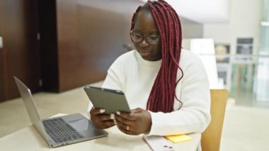 Örgü ören Afrikalı Amerikalı kadın modern ofis ortamında tablet ve laptop kullanıyor..