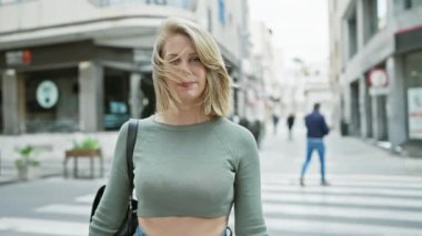 Kısa sarı saçlı, gülümseyen genç bir kadın şehir caddesinde dikilirken kollarını kavuşturuyor, sıradan bir hayat tarzını canlandırıyor..