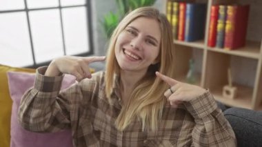 Evdeki neşeli sarışın kadın, koltukta oturuyor, neşeyle işaret ediyor ve diş sağlığını gösteriyor. Güzel bir gülümseme. Dişçiden taze taze.