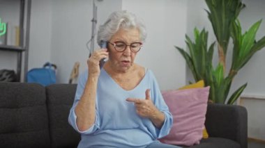 Endişeli, gözlüklü ve gri saçlı yaşlı bir kadın rahat bir oturma odasında akıllı bir telefonla konuşuyor..