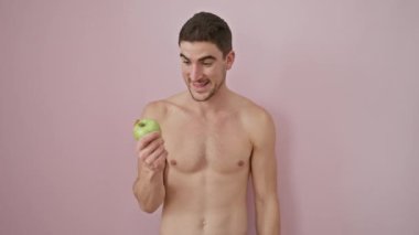 Neşeli, genç İspanyol bir adam, üstsüz, mutlu bir şekilde yukarı bakıyor, eli sağlıklı bir elma tutuyor. İzole edilmiş pembe arka planda geniş bir gülümseme, yan görüntüyü sunuyor. Hoş geldin jesti.!