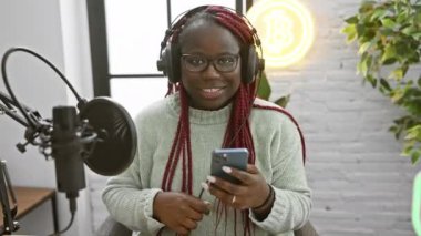 Örgülü, kulaklık takan Afro-Amerikalı bir kadın, arkasında bitcoin logosu olan bir radyo stüdyosunda akıllı telefon kullanıyor..