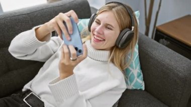 Beyaz kazaklı sarışın kadın evde kulaklıkla koltukta oyun oynuyor..
