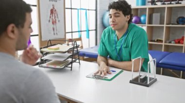 İki adam fizyoterapi kliniğinde, biri önlüklü, biri de terapist olarak parlak bir odada bir hastayla tedaviyi tartışıyor..