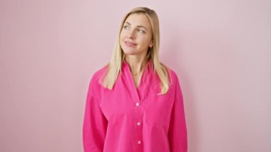 Kendine güveni tam genç bir sarışın kadın rahat, doğal bir poz veriyor, mutlu bir gülümsemeyle başka tarafa bakıyor. Pembe izole arkaplan üzerinde profilde duruyor.