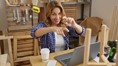 Neşeli genç Latin kadın marangozluk atölyesinde neşe saçıyor! Tahta bir masada oturmuş, kendine güvenen bir gülümsemeyle sana ve kameraya bakıyor, onun güzel ruhunu paylaşıyor.!