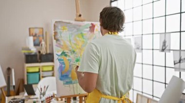 Sarı önlüklü bir adam güneşli bir stüdyoda tuvale resim yapıyor, yaratıcılık ve konsantrasyon sergiliyor..