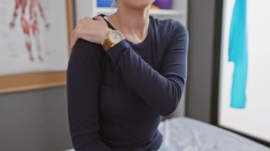 Klinikteki genç bir kadın omzunu anatomik posterlerin önünde acı içinde tutuyor..