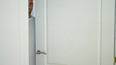 Neşeli bir adam beyaz bir kapıdan içeri bakıyor. Parlak bir evin içinde. Sürpriz, selamlama ve heyecanı resmediyor..