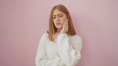 Genç kızıl saçlı kadın diş ağrısı çekiyor, diş hastalıkları elle ağızdan ağıza izole edilmiş pembe arka planda jestler yapıyor.