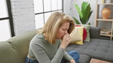 Modern bir oturma odasında yeşil bir kanepede otururken kısa sarı saçlı, endişeli genç bir kadın öksürüyor..