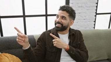 Neşeli genç bir adam, iki eliyle işaret ediyor, evindeki kameraya davetkar bir şekilde bakıyor.