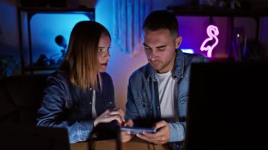 Kot giyen bir adam ve kadın, gece loş bir oyun odasında akıllı bir telefona odaklanmış..