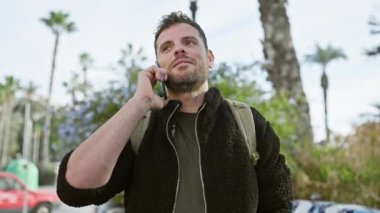 Arka planda palmiye ağaçları olan şehir merkezindeki akıllı telefondan konuşan yakışıklı bir genç..