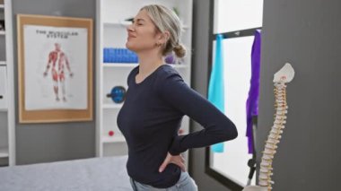 Gündelik kıyafet giymiş genç bir kadın sırt ağrısı çekiyor. Modern bir fizyoterapi kliniğinde duruyor..