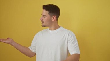 Beyaz tişörtlü, kendinden emin genç İspanyol bir adam. Terfi edip işaret ediyor, sarı izole bir arka plana gülümsüyor, neşe içinde odayı aydınlatıyor..