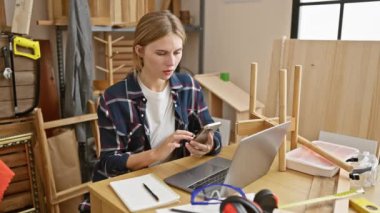 Beyaz saçlı genç bir kadın, ahşap atölyesinde aletleri ve laptopuyla akıllı telefonuyla başarıyı kutluyor..