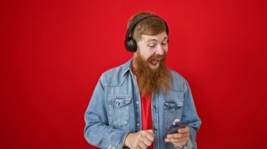 Havalı kızıl saçlı adam heyecanla telefonundan müzik dinliyor, canlı kırmızı bir arka planda kaygısız dans ediyor. Genç adam gülümsüyor, kendine güveni akıyor ve harika melodilerin tadını çıkarıyor.!
