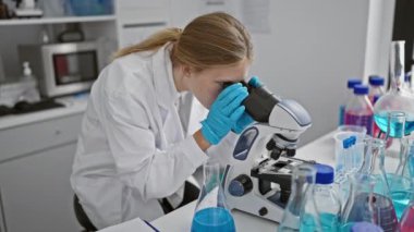 Laboratuvarda çalışan, mikroskop kullanan, beyaz laboratuvar önlüğü ve mavi eldiven giyen neşeli, genç bir bayan bilim adamı..