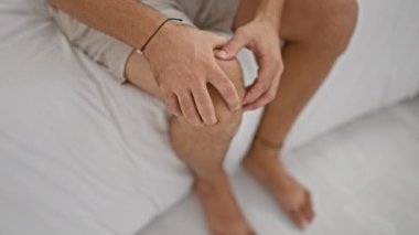 Beyaz yatakta dizine dokunan İspanyol genç adam. Yatak odası iç mekan.