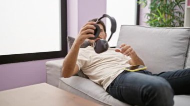 Rahat, Latin kökenli genç bir adam kanepede uzanıyor, rahat bir oturma odasında kulaklıklarıyla akıllı telefonunu kullanıyor..