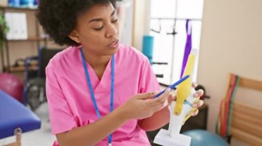 Rehabilitasyon kliniğinde odaklanmış Afrikalı Amerikalı kadın terapist diz modelini değerlendiriyor, sağlık eğitimini sembolize ediyor..