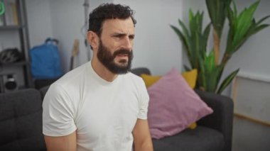 Beyaz tişörtlü orta yaşlı bir adam, açık, şüpheci bir yüzle evde sersemlemiş. İnançsızlığının iğneleyici bakışı şaşkınlık ve şaşkınlığın karışımını ortaya çıkarıyor.