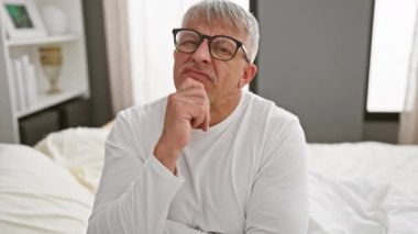 Gergin, orta yaşlı, gri saçlı bir adam yatak odasının sükunetinde endişe verici bir soru soruyor. Eller çenede, pijama kaplı..