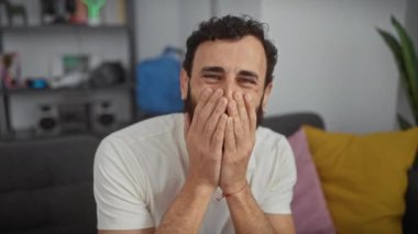 Neşeli orta yaşlı bir adam ev dedikodularından utanıyor, gülümsemesini eliyle örtüyor, beyaz bir tişörtle gülüyor.