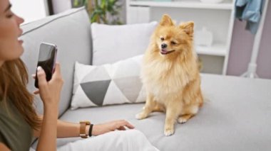 Neşeli genç İspanyol kadın sevgili köpeğiyle koltukta oturuyor, akıllı telefonuyla canlı bir portre çekiyor, evdeki rahat oturma odasında mutluluğu emiyor..