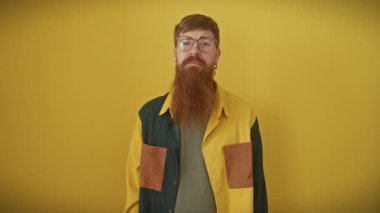 İzole edilmiş sarı bir arka planda duran neşeli kızıl saçlı genç adam gülümsüyor ve el sallıyor. Gözlüklü ve gömlekli kendine güvenen, dostane bir jestle.