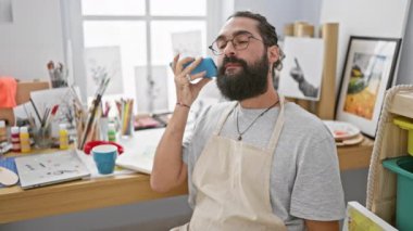 Parlak bir sanat stüdyosunda akıllı telefon kullanan sakallı bir adam. Etrafı boya malzemeleriyle çevrili..