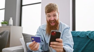 Mutlu kızıl saçlı, kendine güvenen genç adam oturma odasındaki kanepesinden online alışveriş yapmaktan hoşlanıyor. Evde kredi kartıyla ödeme yapmak için telefonunu kullanıyor..