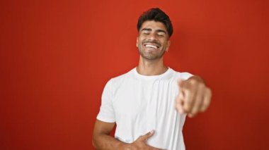 Sakallı, genç bir İspanyol adam kameraya gülümseyip izole edilmiş kırmızı bir arka plana işaret ediyor.