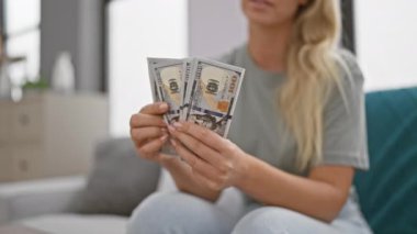 Genç sarışın kadın rahat bir oturma odasında 100 dolarlık banknotlar sayıyor. Finans, tasarruf ve aile hayatı tasviri yapıyor..