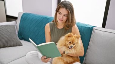 Gülümseyen genç İspanyol kadın evcil köpeğini öpüyor, evinde dinleniyor, koltukta rahatça oturuyor, kitap okuyor, boş zamanların tadını çıkarıyor.