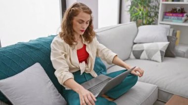 Kanepeye odaklanmış genç bir kadın - dizüstü bilgisayar kullanırken parmak hareketiyle hayır diyor, evde ciddi bir online işe dalmış.