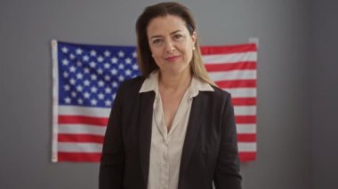 Takım elbiseli kendine güvenen kadın, ofiste Amerikan bayrağıyla kameraya bakıyor.
