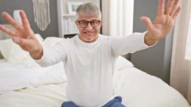 Neşeli, gri saçlı, orta yaşlı, pijama giyen, neşeyle işaret eden, parmaklarıyla sekiz numarayı gösteren bir adam.