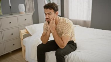 Sakallı, genç bir İspanyol adam modern bir yatak odasında yatağın üzerine oturmuş elleriyle yüzünü kapatıyor..