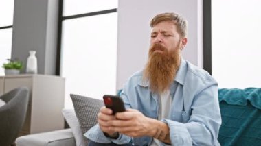 Evde telefonla uğraşırken kanepede boyun ağrısı çeken stresli kızıl saçlı adam.