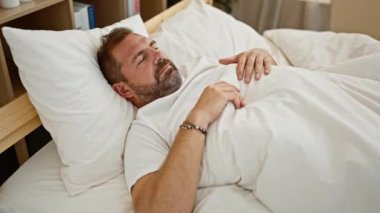 Sakallı ve gri saçlı orta yaşlı bir adam rahat bir yatak odasında huzur içinde uyur. Rahatlık ve rahatlık örneği verir..