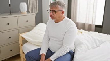 Huysuz orta yaşlı adam, gri saçlı, kuşkucu ve gergin, evde pijama giyiyor onaylamayan yüzüyle, yatak odasında kollarını kavuşturmuş, negatif aura yayıyor..