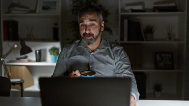 Gri saçlı olgun bir sakallı adam karanlık bir ofiste geç saatlere kadar çalışır, gülümser ve elinde bir belge tutar..