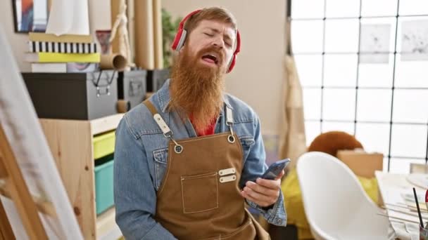 アートスタジオ キャンバス ペイントブラシで音楽を聴きながら踊るハンサム ひげ付き 若い赤毛の男 喜びで微笑む — ストック動画